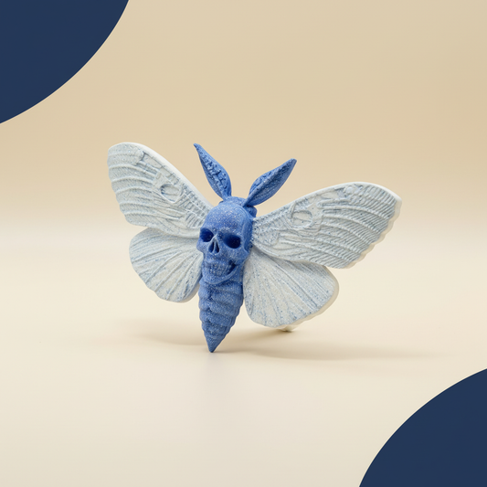Aimant papillon bleu foncé et bleu clair avec tête de mort, ailes blanches, fond crème et ombre naturelle, sans halo lumineux